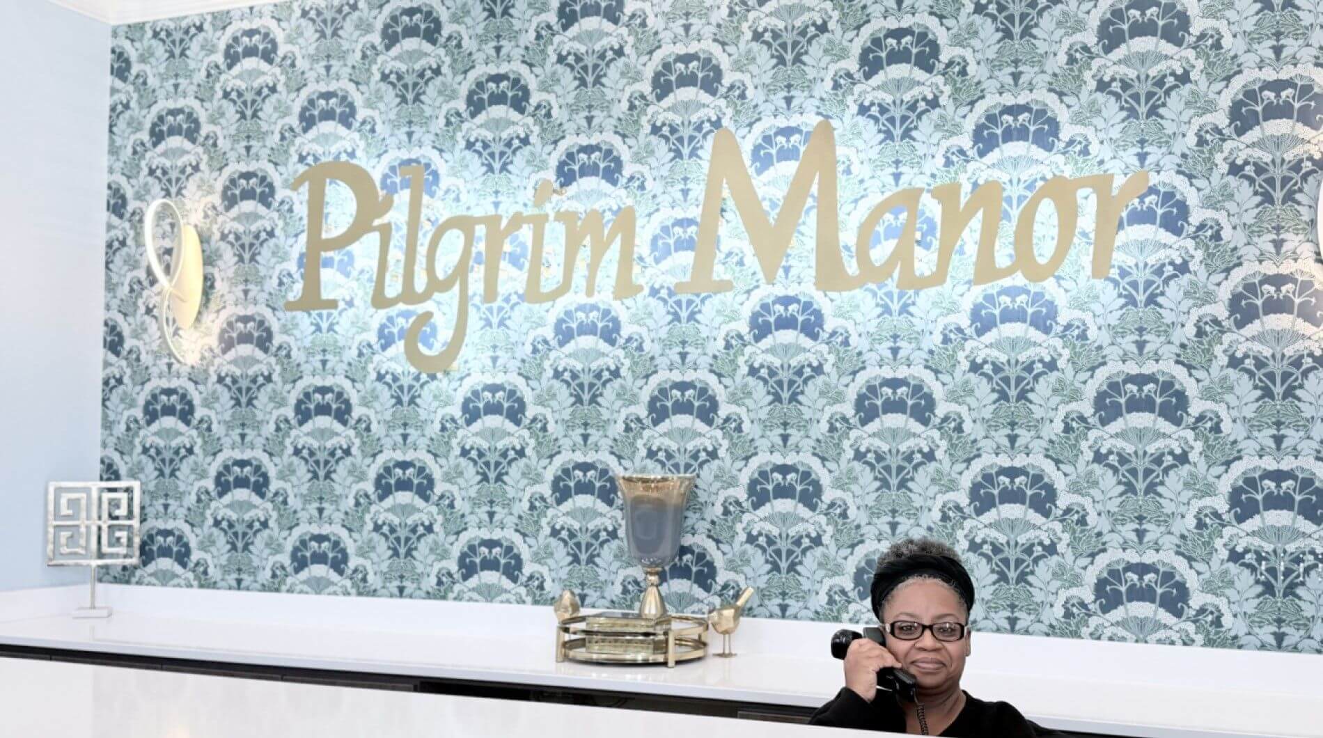 Woman receptionist on phone by Pilgrim Manor sign, highlighting comforts of nursing home care in GreenAcres Place in Bossier City, LA